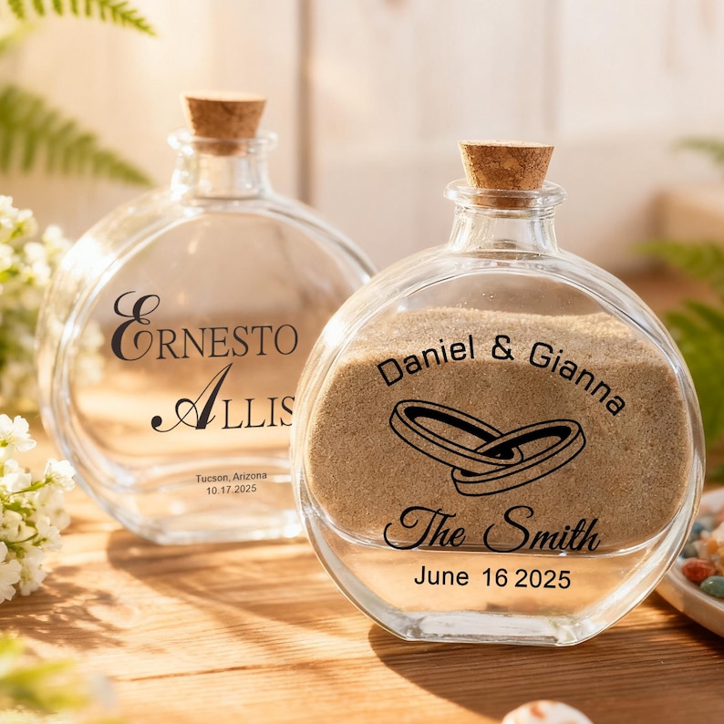 May include: Two round clear glass bottles with cork stoppers. The left bottle features the text Ernesto Allis, Tucson, Arizona, 10.17.2025. The right bottle contains sand and features the text Daniel & Gianna, The Smith, June 16 2025, and two rings.