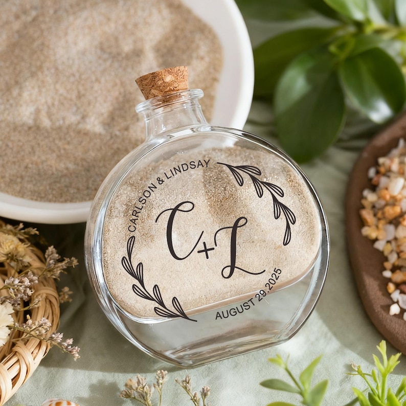 May include: A clear, round glass bottle with a cork stopper, filled with light-colored sand. The front of the bottle features black printed text that reads CARLSON & LINDSAY, C + L, and AUGUST 29, 2025, framed by two delicate black leaf branch illustrations.