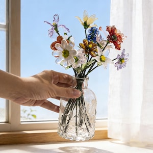 May include: A clear glass vase with an embossed design holds a colorful bouquet of glass flowers. The flowers include white, yellow, red, blue, and purple blooms. The vase sits on a windowsill with a blue sky visible in the background.
