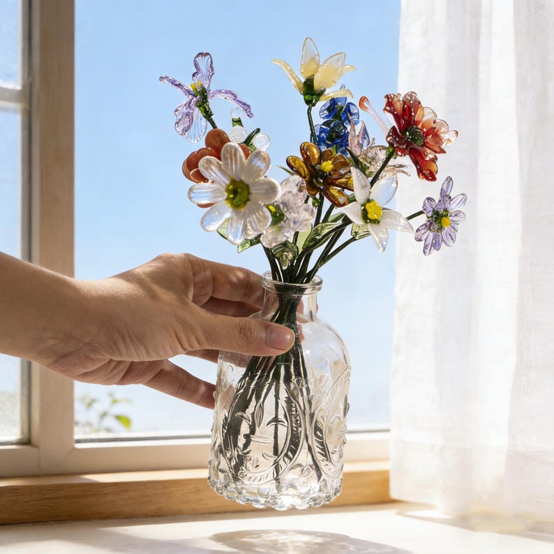 May include: A clear glass vase with an embossed design holds a colorful bouquet of glass flowers. The flowers include white, yellow, red, blue, and purple blooms. The vase sits on a windowsill with a blue sky visible in the background.