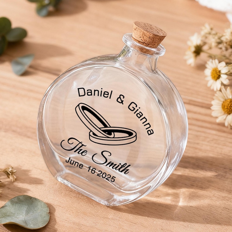 May include: A clear, round glass bottle with a cork stopper. The front features black printed text reading Daniel & Gianna, The Smith, and June 16 2025, along with a graphic illustration of two interlocking wedding rings centered on the glass.