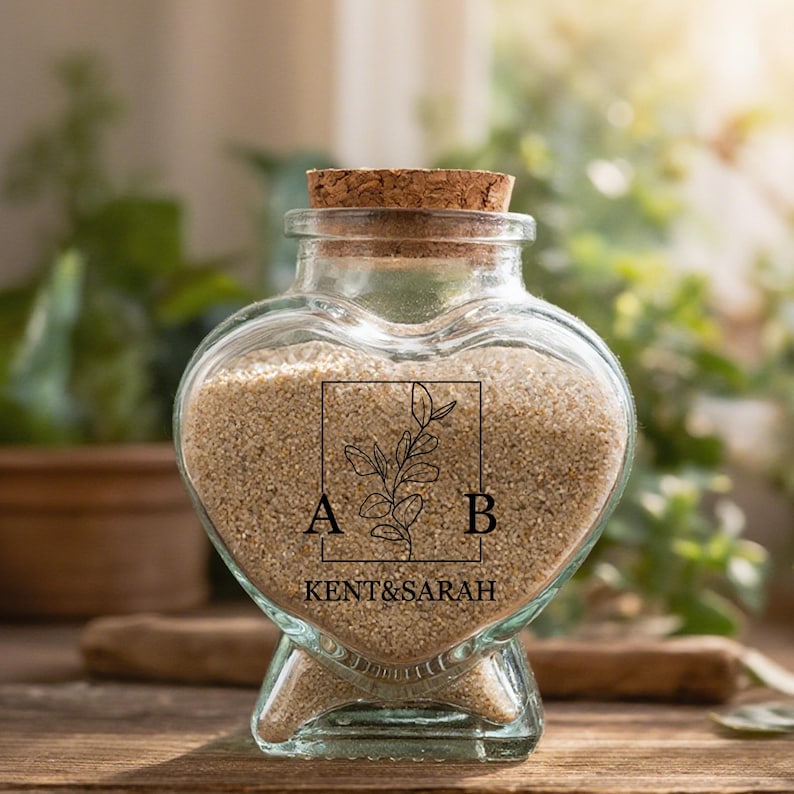 May include: A heart-shaped clear glass jar with a cork stopper, filled with beige sand. The front features a black printed design of a rectangular frame containing a botanical branch illustration, the letters A and B, and the text KENT&SARAH.