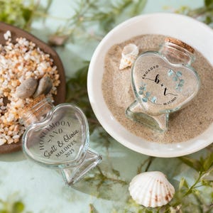 May include: Two heart-shaped glass jars with cork stoppers, filled with sand. One jar features the text 'ENGAGED b + t 06.17.2025' with eucalyptus leaves. The other jar displays 'HONEYMOON SAND Scott & Olivia MALDIVES 2025' in black lettering.