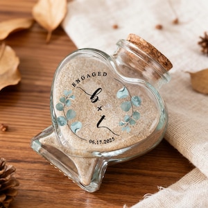 May include: A heart-shaped glass bottle with a cork stopper, filled with light beige sand. The front features a printed design with the word ENGAGED, the letters b + t, and the date 06.17.2025, framed by two watercolor-style eucalyptus branches.
