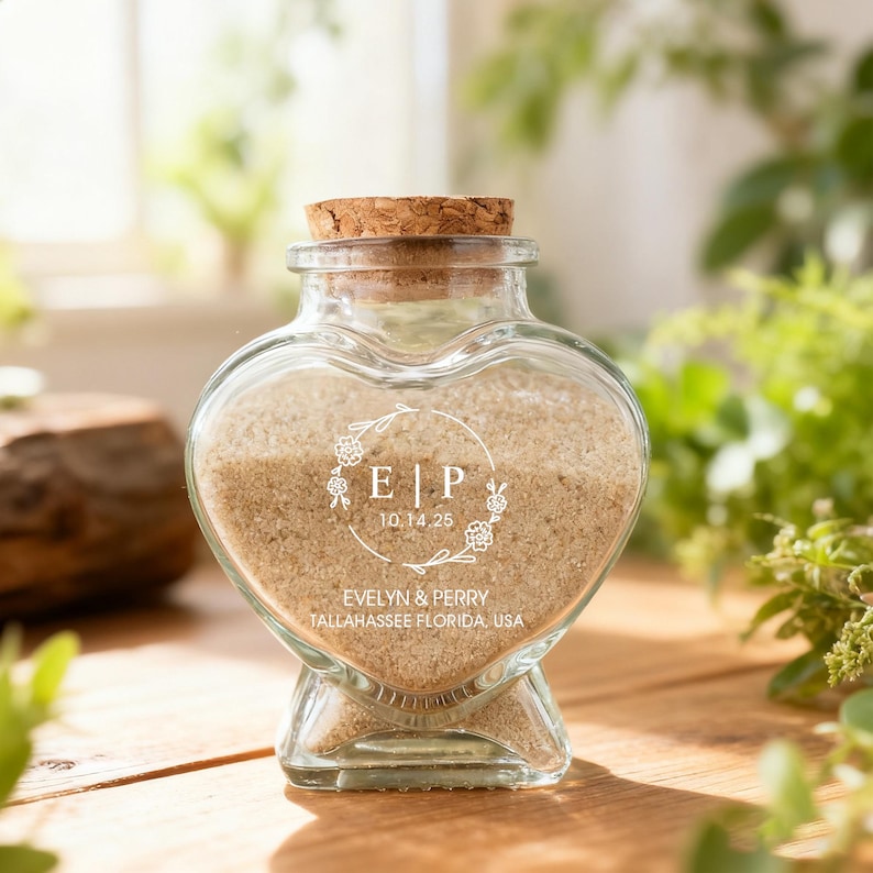 May include: A heart-shaped glass jar with a cork stopper, filled with light tan sand. The front features white printed text reading E | P, the date 10.14.25, and the names EVELYN & PERRY with TALLAHASSEE FLORIDA, USA, framed by a delicate floral wreath.