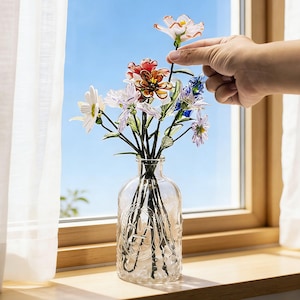 May include: A clear glass vase with a decorative embossed design holds a bouquet of colorful artificial flowers. The flowers include white, red, and blue blooms. The vase sits on a wooden windowsill with a bright blue sky visible through the window.