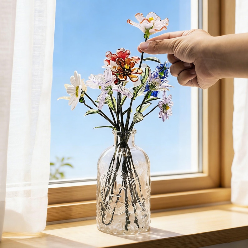 May include: A clear glass vase with a decorative embossed design holds a bouquet of colorful artificial flowers. The flowers include white, red, and blue blooms. The vase sits on a wooden windowsill with a bright blue sky visible through the window.
