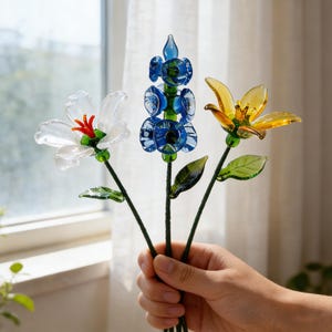 May include: Three glass flowers in a hand. One is white with a red center, one is blue, and the third is yellow. The flowers have green stems and leaves. The background is a window with a white curtain.