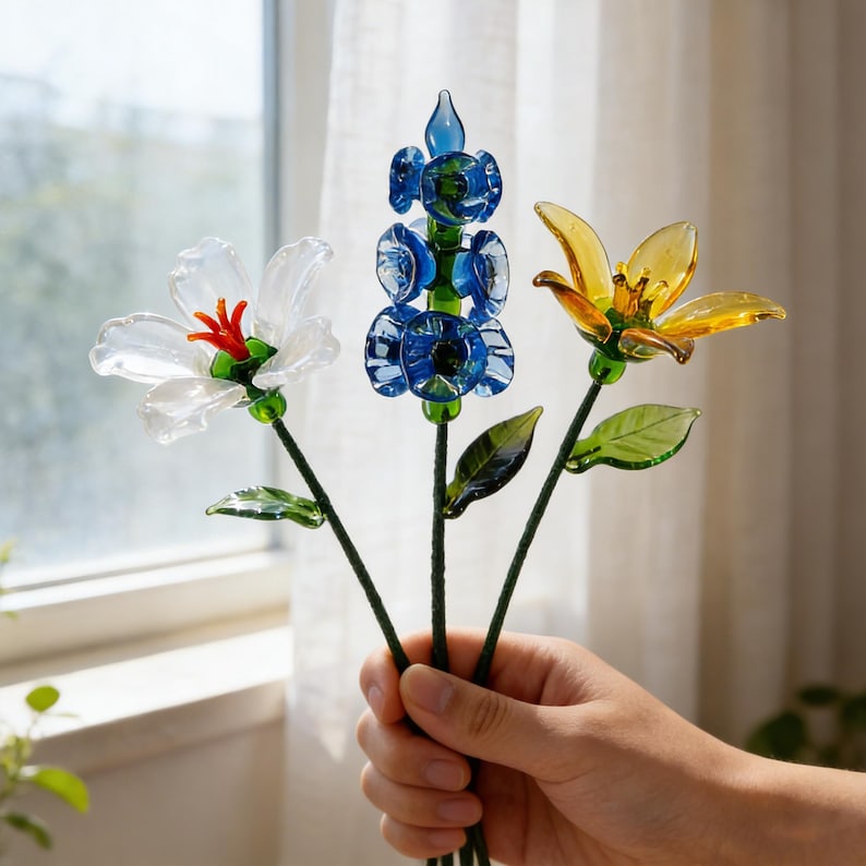 May include: Three glass flowers in a hand. One is white with a red center, one is blue, and the third is yellow. The flowers have green stems and leaves. The background is a window with a white curtain.