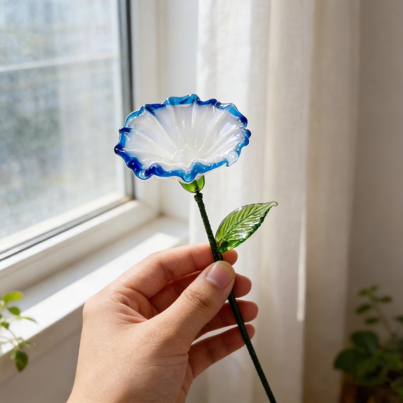May include: A handcrafted glass flower with a white center and a vibrant blue rim. The stem is dark green, with a single green leaf. The flower is held in a hand near a window, suggesting a decorative item or art piece.