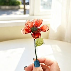 May include: A handcrafted red glass flower with a green stem and leaf. The flower has translucent petals and a dark green center. The stem is held by a person with blue nail polish.