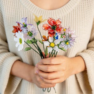 May include: A bouquet of colorful, translucent glass flowers. The arrangement includes red, white, purple, and yellow blooms with green stems. The flowers are held in a person's hands, against a cream-colored sweater.