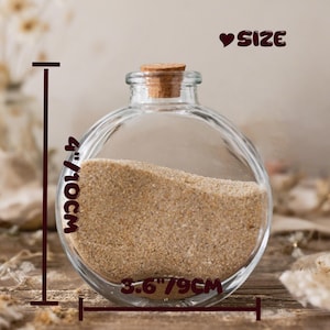 May include: A clear, round glass bottle with a cork stopper, partially filled with beige sand. The text "SIZE" appears at the top right. Measurement lines indicate a height of 4 inches and a width of 3.6 inches for the glass container.