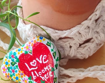May include: A heart-shaped painted rock with a red heart in the center and the words "Love blooms here" written in white. The rock is decorated with colorful flowers and green leaves.