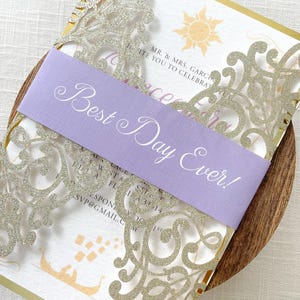 May include: Wedding invitation with a gold border and intricate silver glitter filigree design. A lavender band across the front reads "Best Day Ever!" in elegant script. The invitation includes a sun illustration and event details.