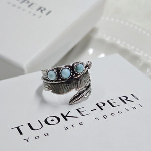 Op de afbeelding: Een zilverkleurige ring in een veervorm, versierd met drie lichtblauwe edelstenen. De ring wordt gepresenteerd op een wit oppervlak met de merknaam "TUOKE-PERI". Het ontwerp van de ring is gedetailleerd.