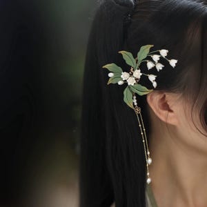 May include: A hair accessory featuring a cluster of white lily of the valley flowers with green leaves and two delicate gold chains dangling with small white pearls. The floral arrangement is attached to a hair clip, creating a botanical design.