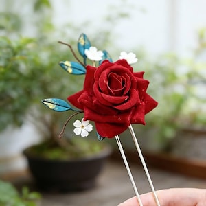 May include: A hair accessory with a large, velvety red rose. The rose is complemented by small white flowers and blue and gold leaves. The hair pin has two long, thin, gold-coloured prongs.