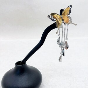 May include: A decorative butterfly hair accessory with gold and silver tones. The butterfly has intricate wing details and is adorned with dangling silver chains and bell-shaped charms. The accessory is mounted on a black display stand.