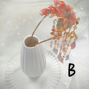 May include: A decorative hair accessory featuring a floral design with red and pink petals, accented with pearls and gold-colored metal. The hairpiece has dangling chains with small, colorful accents. The accessory is displayed in a white vase.