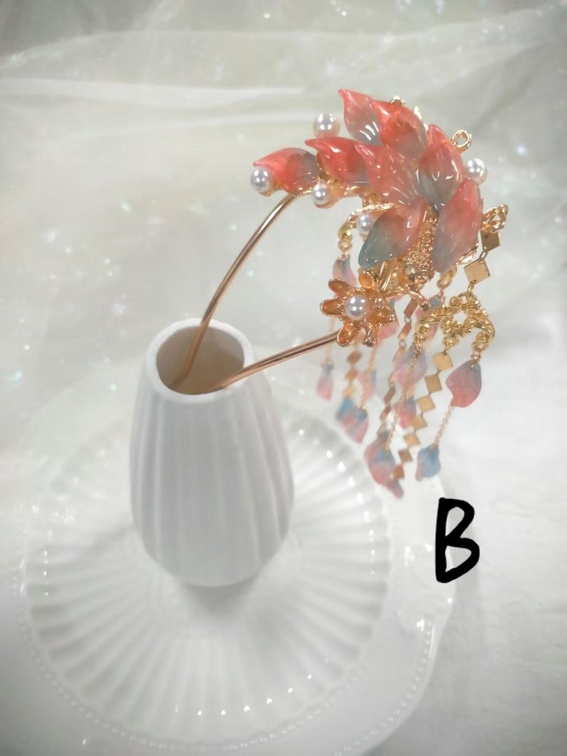 May include: A decorative hair accessory featuring a floral design with red and pink petals, accented with pearls and gold-colored metal. The hairpiece has dangling chains with small, colorful accents. The accessory is displayed in a white vase.