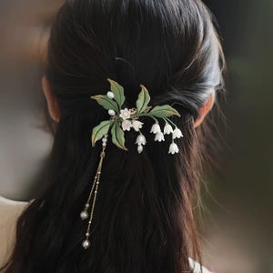 May include: A hair accessory featuring white lily of the valley flowers, green fabric leaves, and small white pearls. The piece includes a delicate gold-toned chain dangling with additional pearls, secured into dark hair to create a floral arrangement.