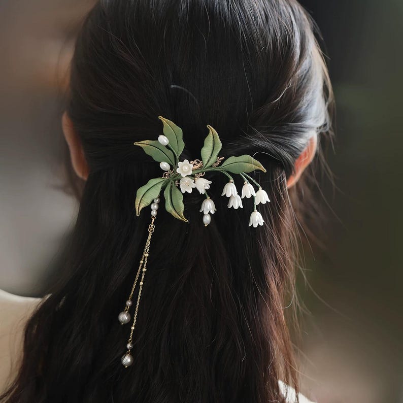 May include: A hair accessory featuring white lily of the valley flowers, green fabric leaves, and small white pearls. The piece includes a delicate gold-toned chain dangling with additional pearls, secured into dark hair to create a floral arrangement.