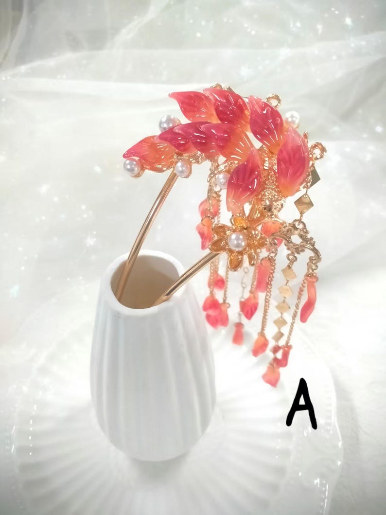 May include: A decorative hair accessory featuring a floral design with red and orange petals, pearls, and gold accents. The hairpiece is held in a white vase. The letter "A" is visible on the plate.