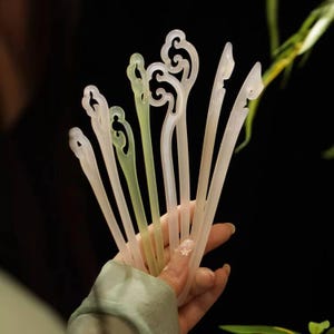 May include: A selection of translucent hair sticks in shades of white and green. Each stick has a decorative, swirling design at the top. The sticks are held together, highlighting their delicate and elegant appearance against a dark background.