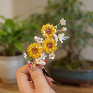 Pode incluir: Um acessório de cabelo floral com três flores amarelas em forma de girassol, centros castanhos, detalhes de pérolas brancas e pequenas flores brancas. Inclui folhas verdes e uma base de arame dourado.