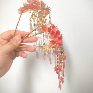 May include: An ornate hair accessory featuring red and orange leaf-shaped accents, pearls, and dangling chains. The gold-toned metal hairpiece is held in a hand against a white background. The design is reminiscent of traditional Asian hair ornaments.
