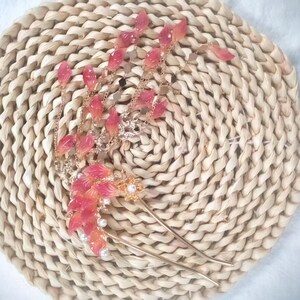 May include: A decorative hair accessory featuring a gold-toned metal design with cascading pink and white leaf-shaped accents. The hair piece includes a pair of long, slender pins and is adorned with small, round pearls. The accessory rests on a woven, circular mat.