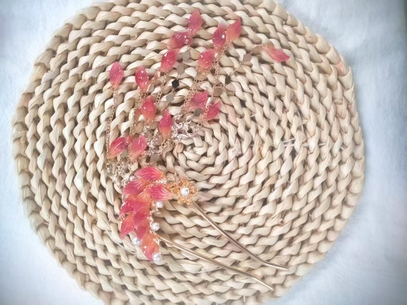 May include: A decorative hair accessory featuring a gold-toned metal design with cascading pink and white leaf-shaped accents. The hair piece includes a pair of long, slender pins and is adorned with small, round pearls. The accessory rests on a woven, circular mat.
