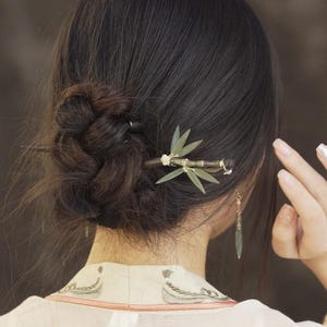 May include: A wooden hair stick with a bamboo leaf design. The hair accessory features green leaves and small white flowers. The hair stick is inserted into a bun of dark brown hair. A gold earring hangs from the ear.