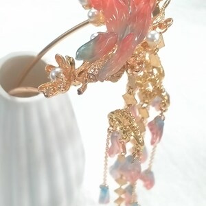 May include: A gold-toned hair accessory featuring a large, ornate floral design with pink, blue, and white accents. The design includes faux pearls and dangling charms, creating an elegant and decorative piece.