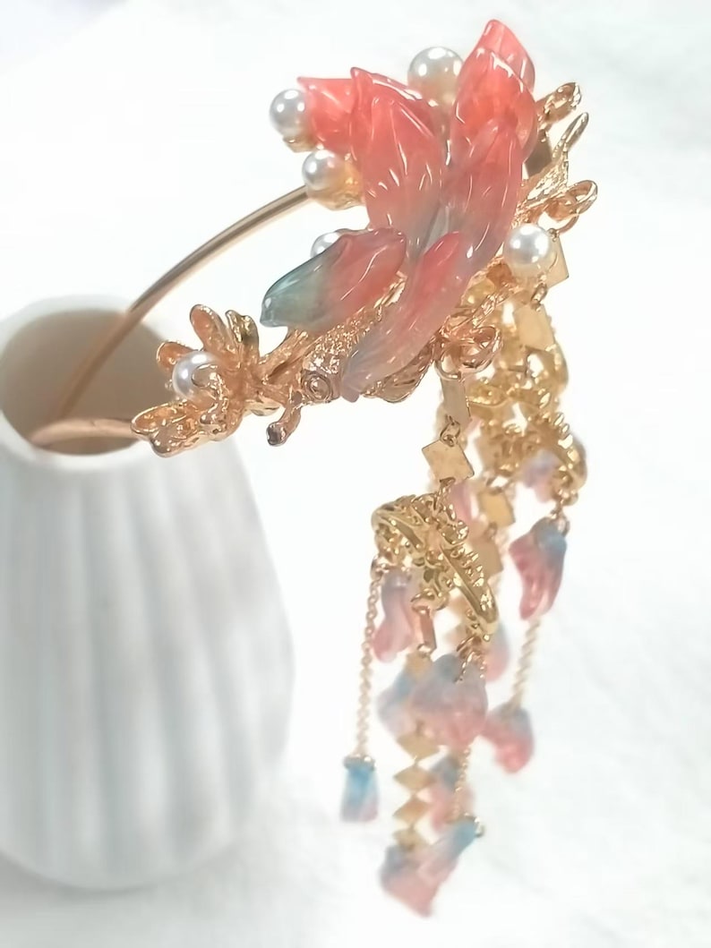 May include: A gold-toned hair accessory featuring a large, ornate floral design with pink, blue, and white accents. The design includes faux pearls and dangling charms, creating an elegant and decorative piece.