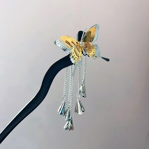 May include: A decorative hair stick featuring a gold and silver butterfly design. The butterfly has intricate detailing and is adorned with a small gem. Silver chains with bell-shaped charms dangle from the butterfly, adding movement and flair. The stick is black.
