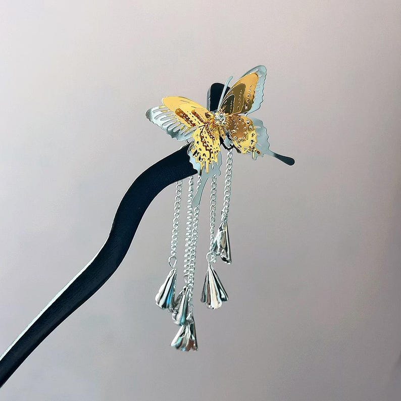May include: A decorative hair stick featuring a gold and silver butterfly design. The butterfly has intricate detailing and is adorned with a small gem. Silver chains with bell-shaped charms dangle from the butterfly, adding movement and flair. The stick is black.