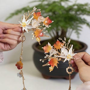 Op de afbeelding: Twee bloemen haarclips met witte, oranje en rode doorschijnende bloemen en goudkleurige accenten. Elke clip heeft een bungelende ketting met decoratieve elementen. Op de achtergrond een plant in een pot.