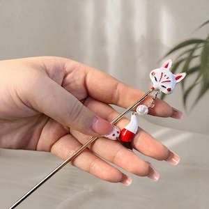 Puede incluir: Un palillo de pelo dorado con una cabeza de zorro blanca con detalles rojos. Un dije blanco y rojo cuelga debajo de la cabeza del zorro, con un pequeño acento de perla. Accesorio para peinado decorativo.
