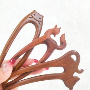 Puede incluir: Una colección de horquillas de madera para el cabello con formas de animales. Las horquillas están talladas en madera marrón oscuro y presentan diseños de gato, zorro y otras figuras estilizadas. Los accesorios para el cabello se mantienen unidos.