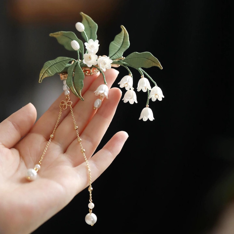 May include: A hair accessory featuring white lily of the valley flowers and green leaves, accented with gold-toned metal chains and dangling white pearls. The floral arrangement is attached to a gold-toned base with delicate metal detailing.