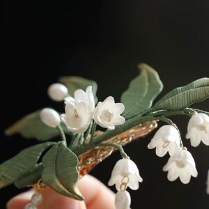 May include: A hair clip featuring a cluster of white bell-shaped lily of the valley flowers and small white pearls. The floral arrangement includes textured green fabric leaves and stems attached to a metallic gold-toned hair clip base.