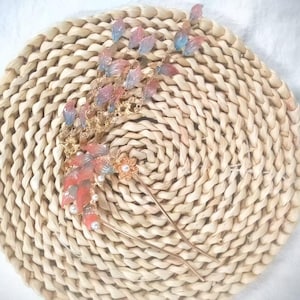 May include: A gold-toned hair accessory with a floral and leaf design. The piece features red, pink, and blue accents, along with small pearls. The accessory is placed on a woven, circular mat.