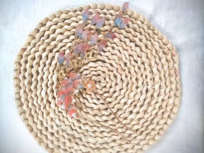 May include: A gold-toned hair accessory with a floral and leaf design. The piece features red, pink, and blue accents, along with small pearls. The accessory is placed on a woven, circular mat.