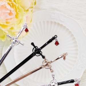 Könnte beinhalten: Nahaufnahme von vier dekorativen Haarnadeln in Schwertform. Die Haarnadeln sind silberfarben, schwarz und goldfarben, jede mit einem roten Edelsteinanhänger. Sie liegen auf einem weißen Teller.