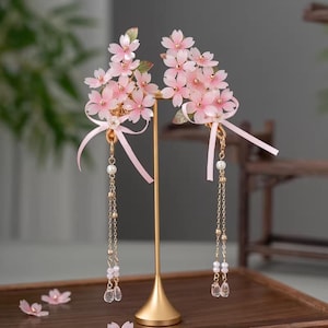 Könnte beinhalten: Zwei rosafarbene Haaraccessoires mit floralem Muster, goldenen Stielen und hängenden Ketten. Jedes Accessoire zeigt Gruppen von rosa Blüten, grüne Blätter und zarte Bänder. Die Ketten sind mit Perlen und klaren, facettierten Perlen verziert.