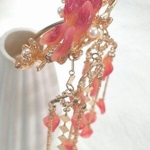 May include: A decorative hair accessory featuring a floral design with pink and orange petals, faux pearls, and gold-colored metal accents. Dangling chains with leaf-shaped charms add movement to this elegant piece.