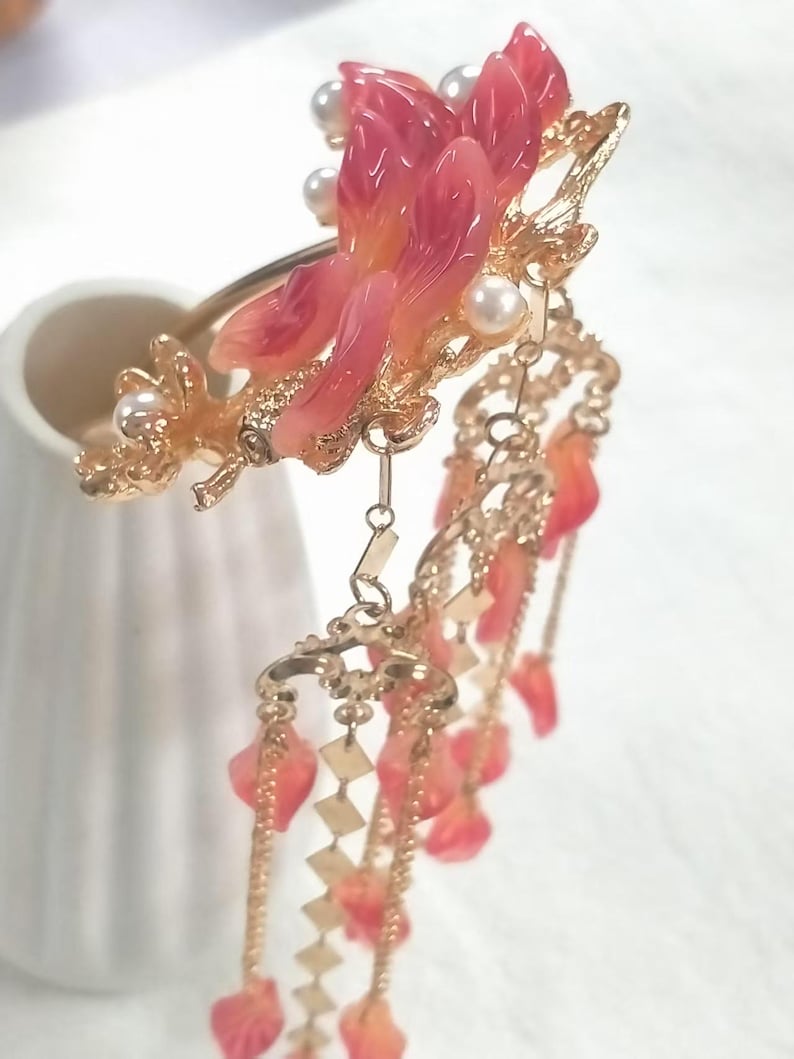 May include: A decorative hair accessory featuring a floral design with pink and orange petals, faux pearls, and gold-colored metal accents. Dangling chains with leaf-shaped charms add movement to this elegant piece.