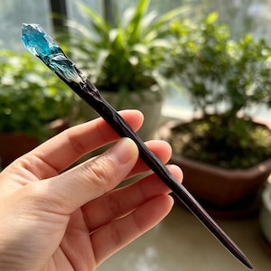Pode incluir: Um palito de cabelo com uma haste de madeira longa, castanho-escura e cónica, rematada com uma peça translúcida, irregular e semelhante a um cristal em tons de azul profundo e branco, criando um contraste com a madeira natural.